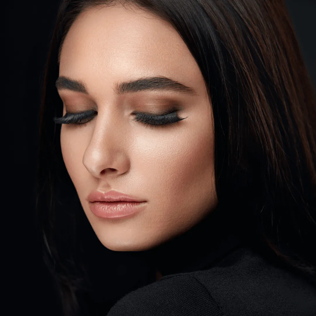 Close-up of a woman with full lash extensions, eyes closed, against black